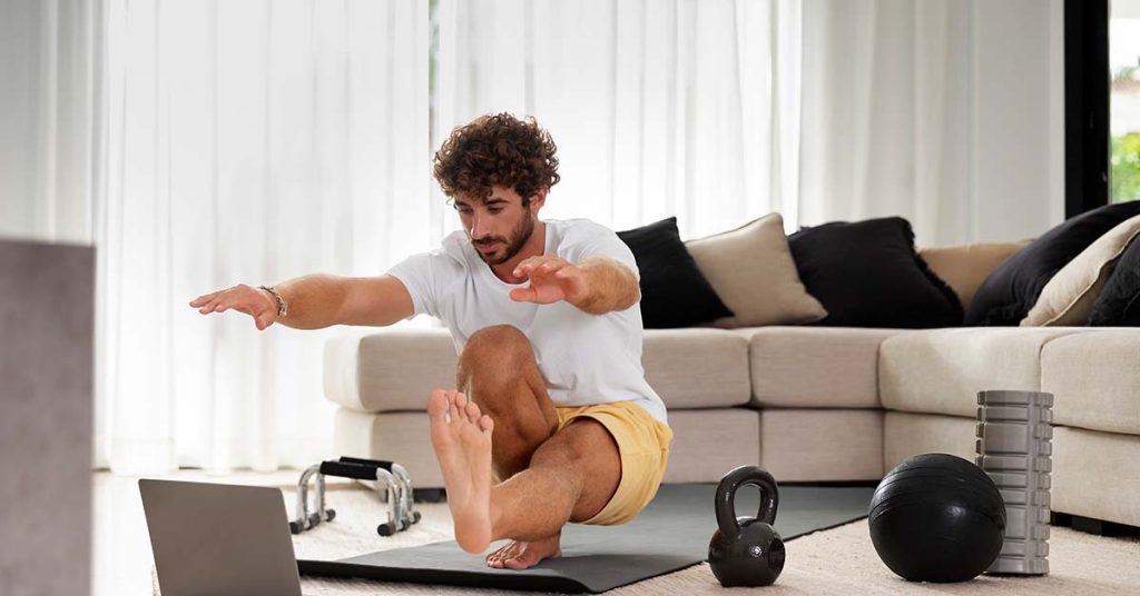 A man performing a one-legged squat in his living room while following a virtual personal training session on his laptop.